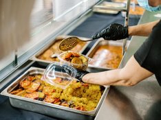 Aus einer großen Metallschale mit Essen nimmt eine Person mit Handschuhen eine Kelle und füllt sie in ein kleines Plastikgefäß
