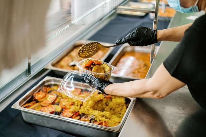 Aus einer großen Metallschale mit Essen nimmt eine Person mit Handschuhen eine Kelle und füllt sie in ein kleines Plastikgefäß