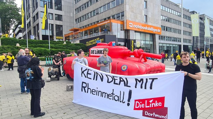 Ein roter aufblaspanzer, davor Menschen mit einem Transpi "Kein Deal mit Rheinmetall"